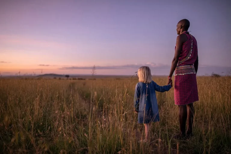 Family tour- a masai moran and a young girl holds hands in the Mara savannah at sunset